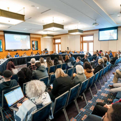 A U‑shaped panel of about a dozen people seated at a long table conducts a formal meeting or board session in a conference room, with two projection screens showing slides at the front. Dozens of attendees sit in rows facing the panel.